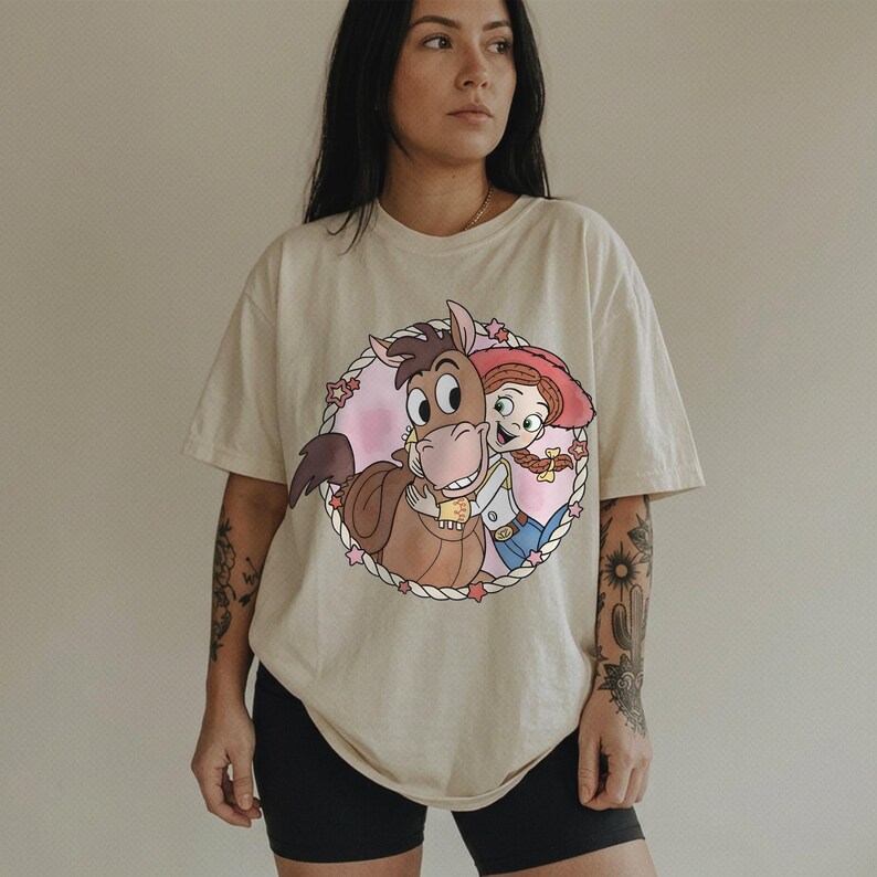 May include: Cream-colored t-shirt featuring a cartoon illustration of a horse hugging a cowgirl. The graphic is framed by a pink circle with stars and a rope border. The cowgirl wears a red hat, blue jeans, and a white shirt.