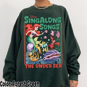 Pu&ograve; includere: Felpa verde foresta con una grafica colorata di "Sing Along Songs" della Disney, con Ariel la sirena, pesci e un granchio. Il testo "THE UNDER SEA" &egrave; sotto la grafica. La felpa &egrave; a girocollo.