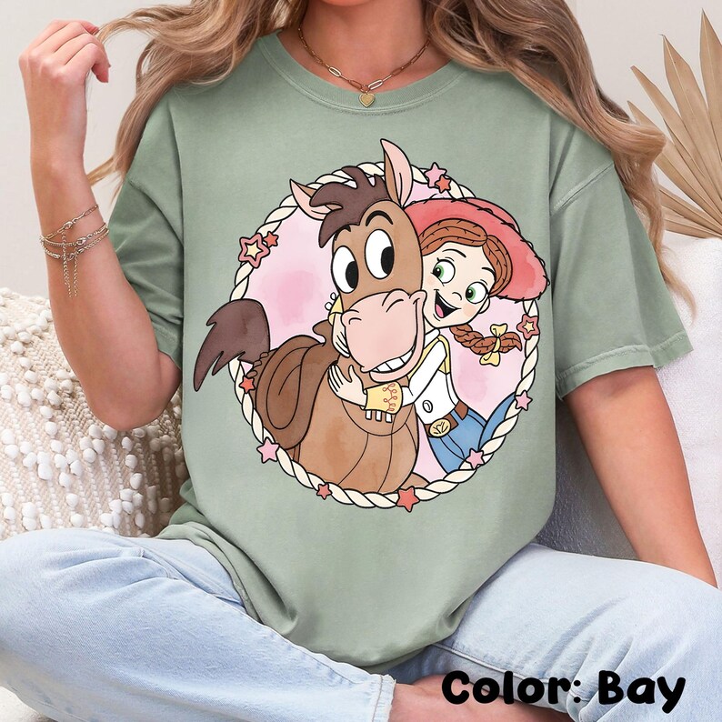 May include: A light green t-shirt featuring a cartoon illustration of a horse hugging a cowgirl inside a pink circle with stars. The text "Color: Bay" is at the bottom of the shirt.