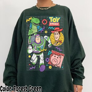May include: Forest green sweatshirt featuring a colorful graphic of characters from the animated film "Toy Story." The design includes Buzz Lightyear, Woody, Rex, and Hamm, with the words "Toy Story" in a retro font. The sweatshirt has long sleeves.