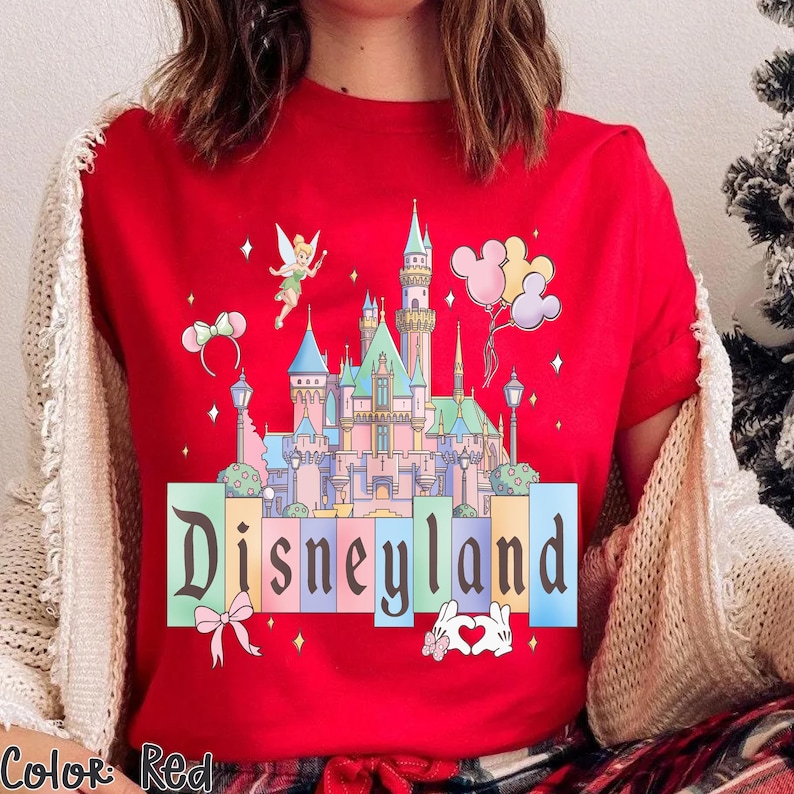 Puede incluir: Camiseta roja con un dise&ntilde;o del castillo de Disneyland, globos de Mickey Mouse y la palabra "Disneyland". El dise&ntilde;o incluye una hada, un lazo, orejas de rat&oacute;n y un gesto de mano en forma de coraz&oacute;n.