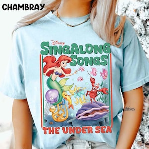 Pu&ograve; includere: T-shirt azzurra con una grafica colorata della serie "Sing Along Songs" di Disney. Il design include Ariel la sirena, varie creature marine e il testo "THE UNDER SEA". La maglietta ha maniche corte.