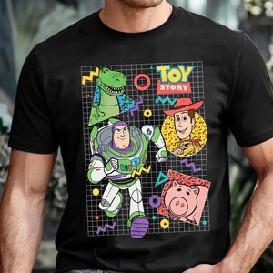 May include: Black t-shirt featuring a colorful graphic of characters from the movie "Toy Story." The design includes Buzz Lightyear, Woody, Rex, and Hamm, set against a grid background with the words "Toy Story."