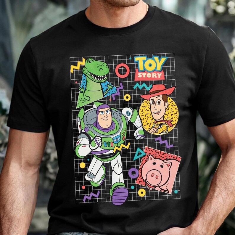 May include: Black t-shirt featuring a colorful graphic of characters from the movie "Toy Story." The design includes Buzz Lightyear, Woody, Rex, and Hamm, set against a grid background with the words "Toy Story."
