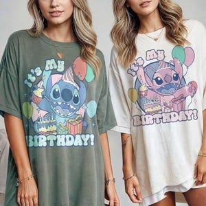 May include: Two oversized t-shirts, one olive green and one off-white, each featuring a cartoon character with the text "It's My Birthday!". The graphic includes balloons, a birthday cake, and a gift box. The shirts are short-sleeved.