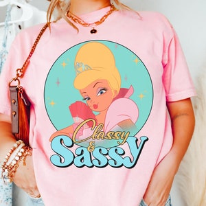 May include: Pale pink t-shirt with a cartoon image of a woman with blonde hair, a tiara, and pink clothing, set against a teal circle with stars. The words "Classy & Sassy" are written in a retro font below the illustration.