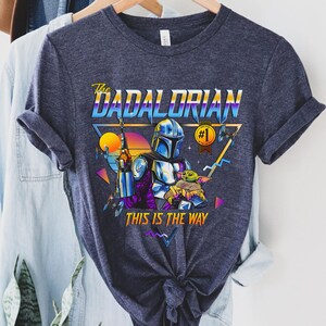 May include: A navy blue t-shirt with a retro-style graphic of a Mandalorian character holding a child. The text "The Dadalorian #1 This is the Way" is printed on the shirt.