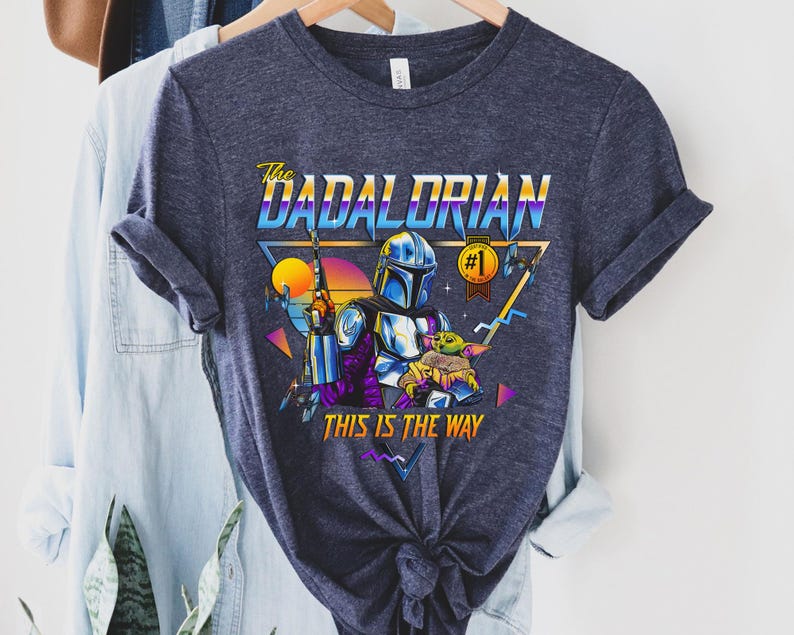 May include: A navy blue t-shirt with a retro-style graphic of a Mandalorian character holding a child. The text "The Dadalorian #1 This is the Way" is printed on the shirt.