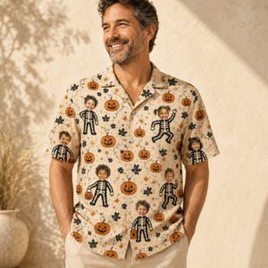 Op de afbeelding: Een beige overhemd met korte mouwen en knoopsluiting, voorzien van een all-over print met oranje pompoenen, zwarte esdoornbladeren en herhalende illustraties van kinderen in skeletkostuums met een Halloween-thema.