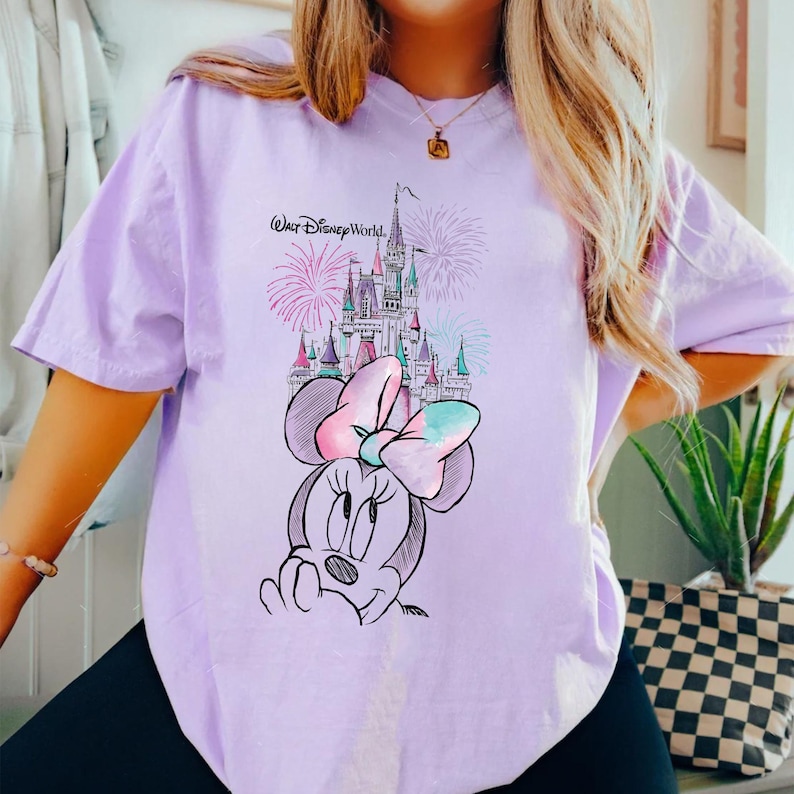Pode incluir: Camiseta lil&aacute;s claro com uma ilustra&ccedil;&atilde;o da Minnie Mouse, um la&ccedil;o e um castelo. O texto "Walt Disney World" est&aacute; acima do castelo. Fogos de artif&iacute;cio no fundo.