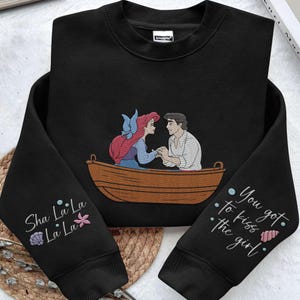 May include: Black sweatshirt featuring a graphic of Ariel and Prince Eric from The Little Mermaid in a boat. The text "Sha La La La La La" is on one sleeve and "You got to kiss the girl" is on the other sleeve.