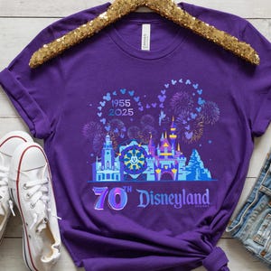 May include: Purple t-shirt with a graphic of the Disneyland castle and fireworks. The text "1955 2025" and "70th Disneyland Resort" is printed on the shirt.