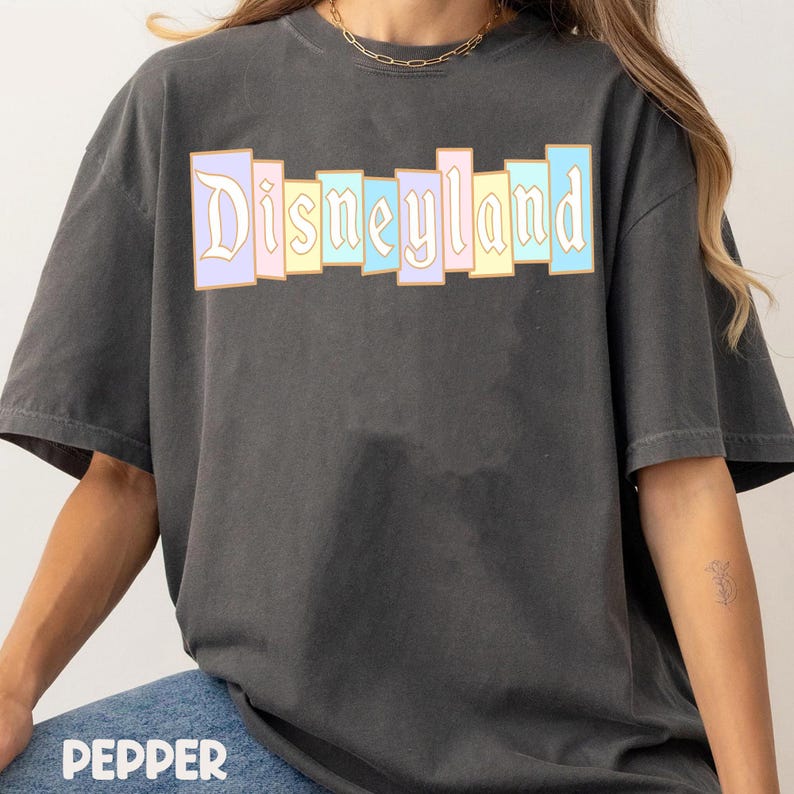 Puede incluir: Una camiseta gris oscuro con la palabra "Disneyland" en colores pastel. El texto est&aacute; dispuesto en un dise&ntilde;o de bloques verticales. La camiseta tiene mangas cortas y un corte relajado. La palabra "Pepper" est&aacute; impresa en la parte inferior.