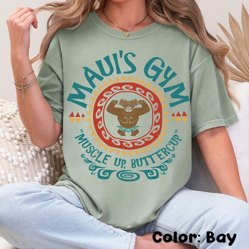 May include: Bay-colored t-shirt with the text "MAUI'S GYM" in teal. A brown cartoon figure flexes inside a red and yellow sun design. The phrase "MUSCLE UP, BUTTERCUP" is below the graphic. The shirt is a relaxed fit.