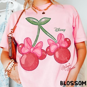Op de afbeelding: Roze T-shirt met een kersontwerp met Minnie Mouse-strikken en het Disney-logo. De kersen zijn rood met roze strikken en groene stelen. De tekst "Minnie Mouse" en "BLOSSOM" staan ook op het T-shirt.