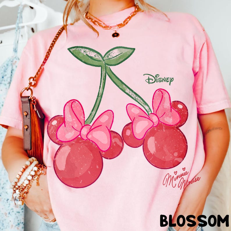 Op de afbeelding: Roze T-shirt met een kersontwerp met Minnie Mouse-strikken en het Disney-logo. De kersen zijn rood met roze strikken en groene stelen. De tekst "Minnie Mouse" en "BLOSSOM" staan ook op het T-shirt.