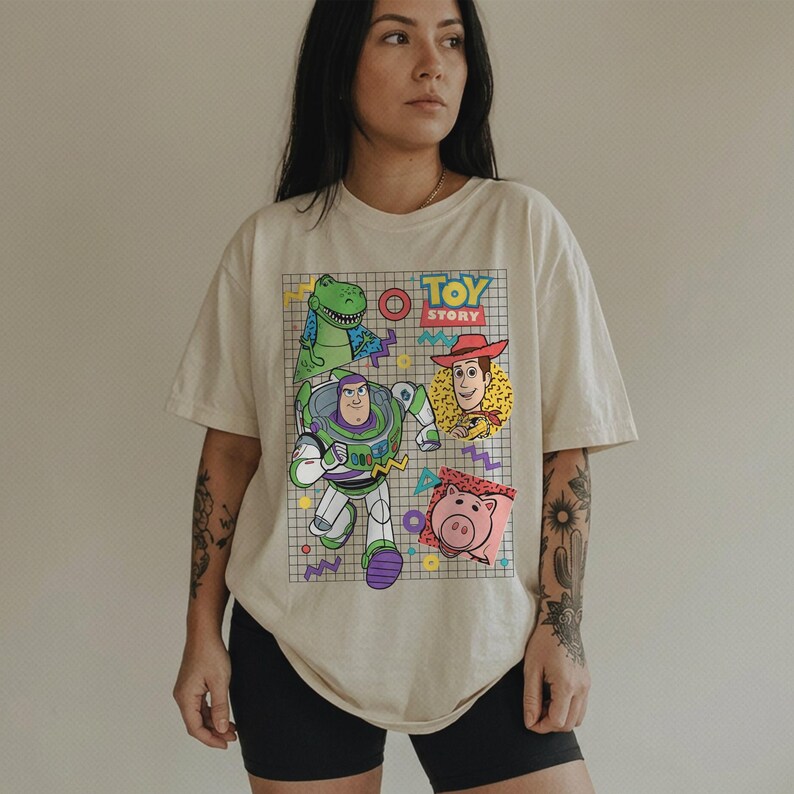 May include: Cream-colored t-shirt featuring a graphic of Toy Story characters: Rex, Buzz Lightyear, Woody, and Hamm. The design includes the words "TOY STORY" in a retro font, set against a grid pattern with colorful accents.