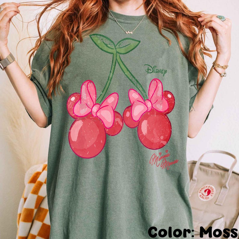 Op de afbeelding: Een mosgroen t-shirt met een Minnie Mouse kersontwerp. De afbeelding toont twee rode kersen met roze strikken, groene stelen en de Disney- en Minnie Mouse-logo's. De kleur van het shirt is 'Moss'.