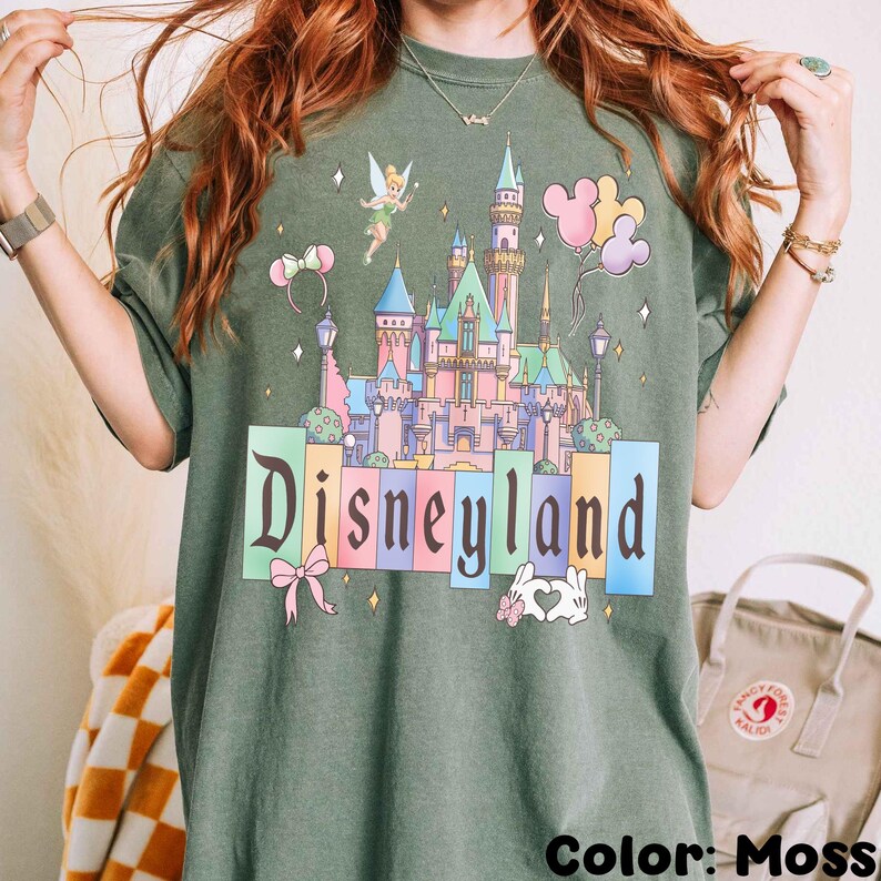 Puede incluir: Camiseta verde musgo con un castillo de Disneylandia colorido, Campanilla, globos de Mickey Mouse y la palabra "Disneyland". El dise&ntilde;o incluye un lazo y un conejito con un coraz&oacute;n.