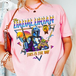 May include: Pink t-shirt with a retro-style graphic of a Mandalorian character with a baby Yoda. The graphic includes the text "The Dadalorian" and "This is the Way".