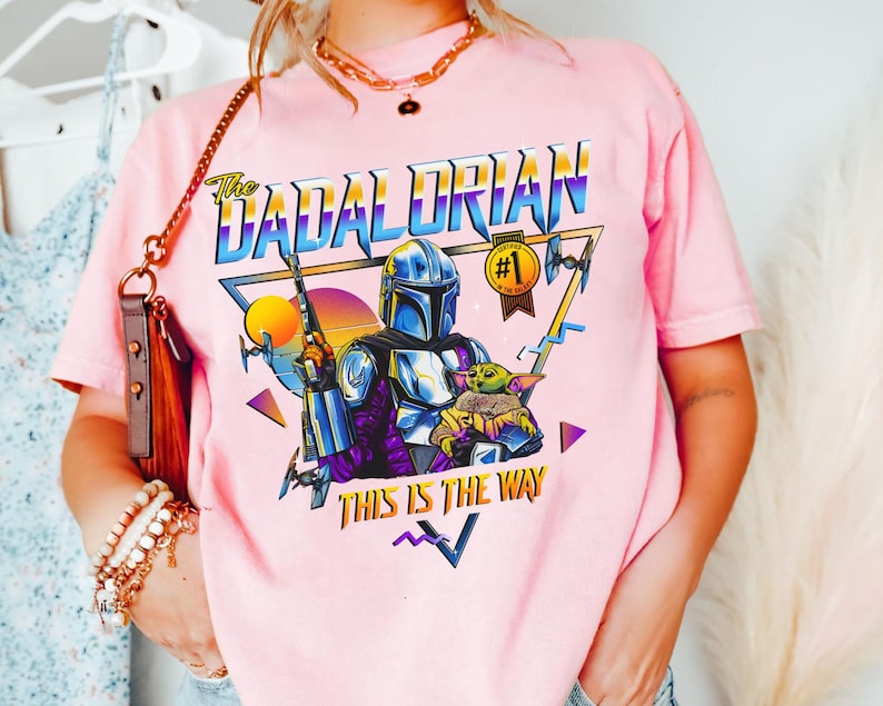 May include: Pink t-shirt with a retro-style graphic of a Mandalorian character with a baby Yoda. The graphic includes the text "The Dadalorian" and "This is the Way".