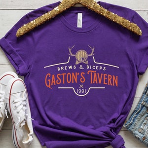 Op de afbeelding: Paars T-shirt met een wit grafisch ontwerp met de tekst "Brews & Biceps Gaston's Tavern 1991". De afbeelding toont een cartoon van een bierpul met geweien.