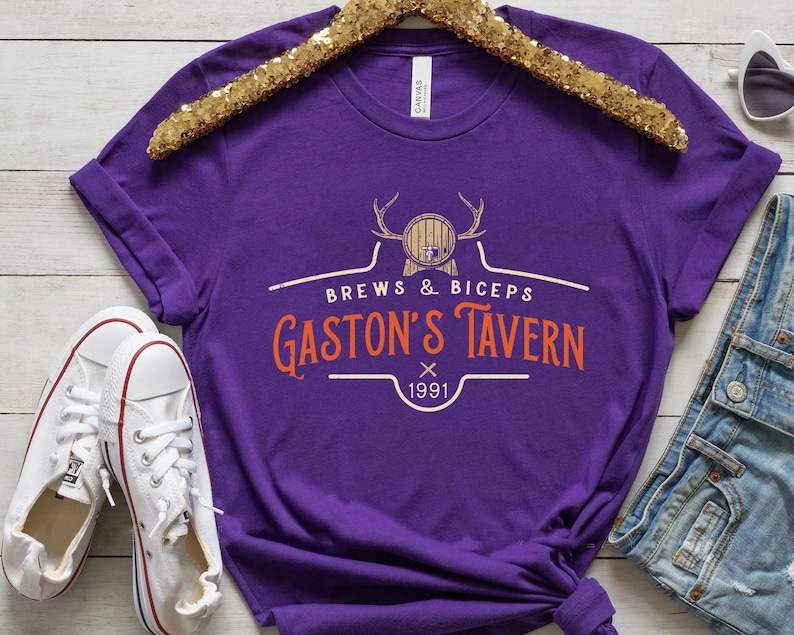 Op de afbeelding: Paars T-shirt met een wit grafisch ontwerp met de tekst "Brews & Biceps Gaston's Tavern 1991". De afbeelding toont een cartoon van een bierpul met geweien.