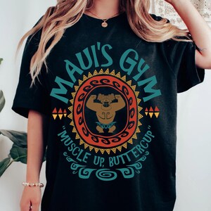 May include: Black t-shirt with a circular design featuring the text "MAUI'S GYM" in teal. The design includes a muscular figure and the phrase "MUSCLE UP, BUTTERCUP" in a decorative font. The shirt is a crew neck.