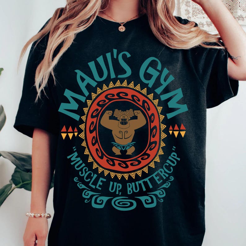 May include: Black t-shirt with a circular design featuring the text "MAUI'S GYM" in teal. The design includes a muscular figure and the phrase "MUSCLE UP, BUTTERCUP" in a decorative font. The shirt is a crew neck.