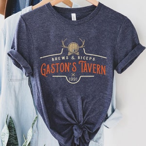 Op de afbeelding: Een marineblauw t-shirt met een vintage-stijl logo met de tekst "Brews & Biceps Gaston's Tavern 1991". Het logo toont een cartoonhert met geweien en een vat.