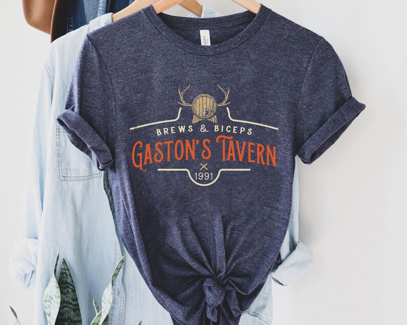 Op de afbeelding: Een marineblauw t-shirt met een vintage-stijl logo met de tekst "Brews & Biceps Gaston's Tavern 1991". Het logo toont een cartoonhert met geweien en een vat.