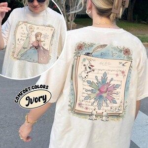 May include: Ivory-colored t-shirt featuring a colorful illustration of characters from a popular animated movie. The front of the shirt displays two characters, while the back showcases sheet music, a snowflake, and the phrase "Let It Go."