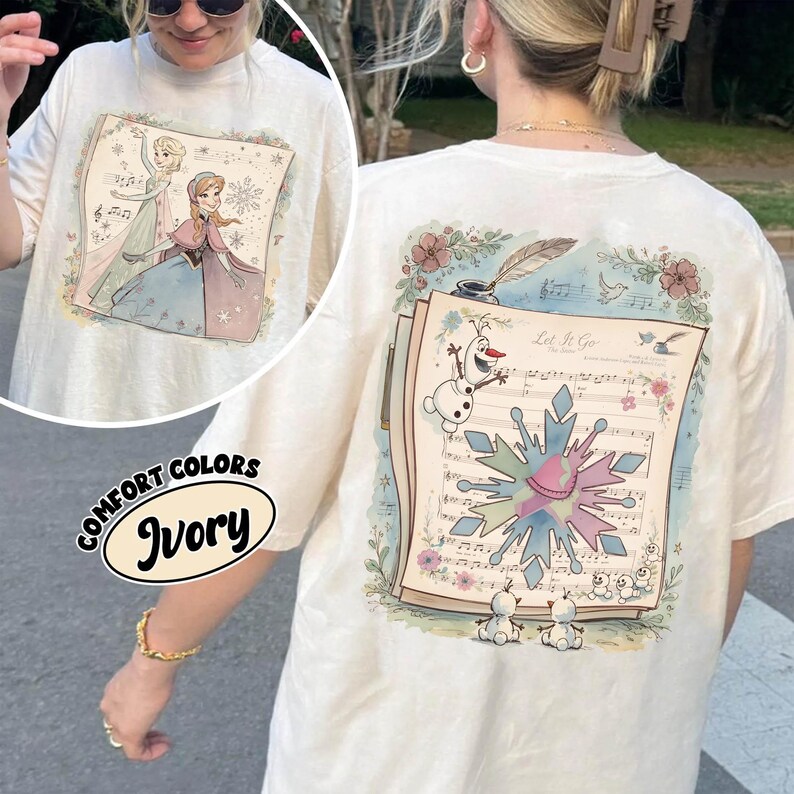May include: Ivory-colored t-shirt featuring a colorful illustration of characters from a popular animated movie. The front of the shirt displays two characters, while the back showcases sheet music, a snowflake, and the phrase "Let It Go."