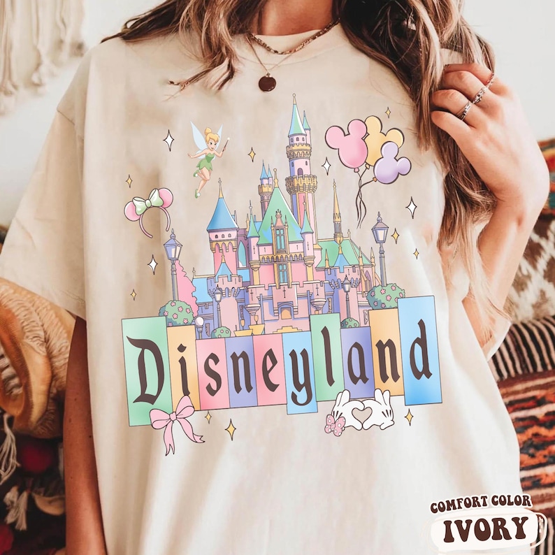 Puede incluir: Camiseta color marfil con un gr&aacute;fico colorido de Disneyland. El dise&ntilde;o incluye un castillo, Campanilla, globos de Mickey Mouse y la palabra "Disneyland". La camiseta est&aacute; etiquetada como "Comfort Color Ivory".