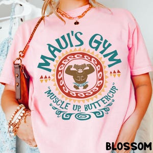 May include: Pink t-shirt with the text "Maui's Gym" in teal and a graphic of a muscular figure. The shirt also features the phrase "Muscle Up, Buttercup" in teal. The word "Blossom" is printed at the bottom.