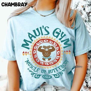 May include: Light blue t-shirt with the text "MAUI'S GYM" in teal and a graphic of a muscular figure. The design includes a red and orange circular emblem with the phrase "MUSCLE UP, BUTTERCUP". The word "CHAMBRAY" is at the top.