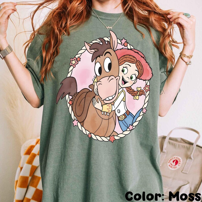 May include: Moss green t-shirt featuring a cartoon graphic of a horse hugging a cowgirl. The graphic is framed by a rope-like border with pink stars. The text "Color: Moss" is at the bottom.