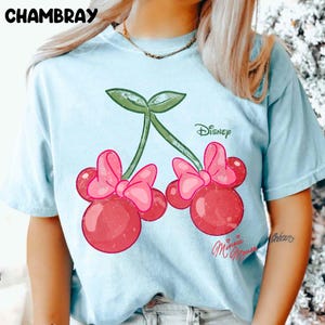 Op de afbeelding: Lichtblauw T-shirt met een kersendesign met Minnie Mouse strikken. De kersen zijn rood met roze strikken en groene stelen. Het woord "Disney" staat boven de kersen en "Minnie Mouse" staat eronder.