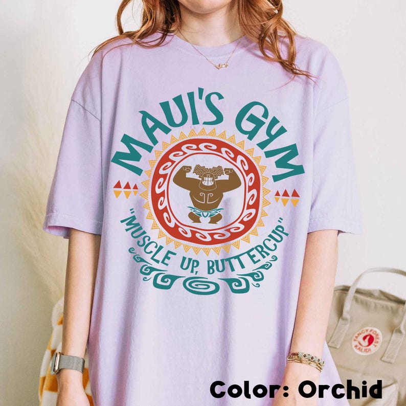 May include: Orchid-colored t-shirt with a graphic that reads "Maui's Gym" in teal lettering. The design includes a cartoon figure flexing inside a red and orange circle, with the text "Muscle Up, Buttercup" below. The shirt is a relaxed fit.