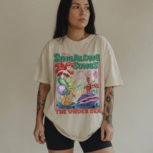 Pu&ograve; includere: T-shirt color crema con una grafica colorata della serie "Sing Along Songs" di Disney. Il design include Ariel la sirena, un granchio e altre creature marine. Il testo "THE UNDER SEA" &egrave; sotto la grafica.