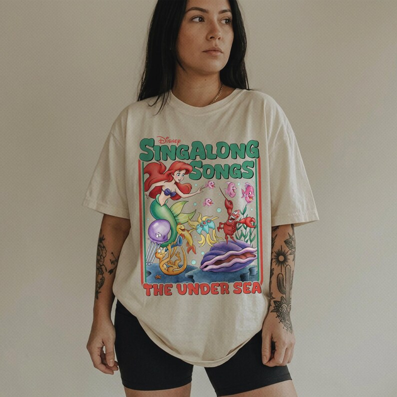 Pu&ograve; includere: T-shirt color crema con una grafica colorata della serie "Sing Along Songs" di Disney. Il design include Ariel la sirena, un granchio e altre creature marine. Il testo "THE UNDER SEA" &egrave; sotto la grafica.