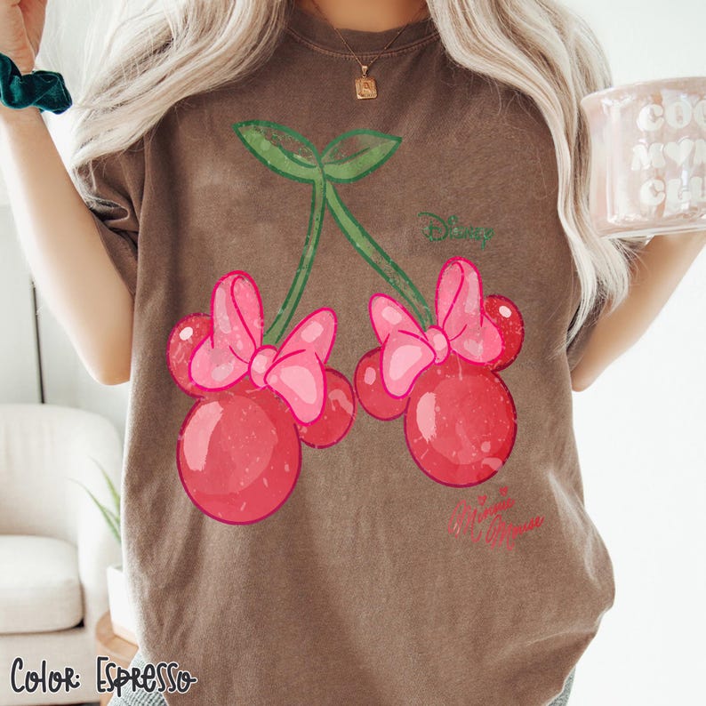 Op de afbeelding: Espresso-kleurig T-shirt met een grillig kersendesign. De kersen zijn rood met roze strikken en groene stelen. Het woord "Disney" is groen gedrukt en "Minnie Mouse" roze.