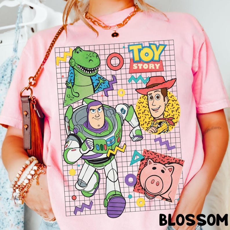 May include: Pink t-shirt featuring a colorful graphic of characters from the animated film "Toy Story." The design includes Buzz Lightyear, Woody, Rex, and Hamm, set against a grid background with the words "Toy Story."