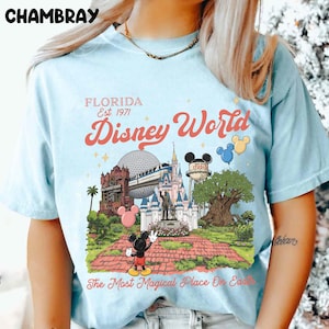 Puede incluir: Camiseta azul claro con un gr&aacute;fico colorido de Disney World. El dise&ntilde;o incluye el texto "FLORIDA Est. 1971" y "Disney World", junto con ilustraciones de caracter&iacute;sticas ic&oacute;nicas del parque y Mickey Mouse. La camiseta tambi&eacute;n tiene el texto "The Most Magical Place On Earth".