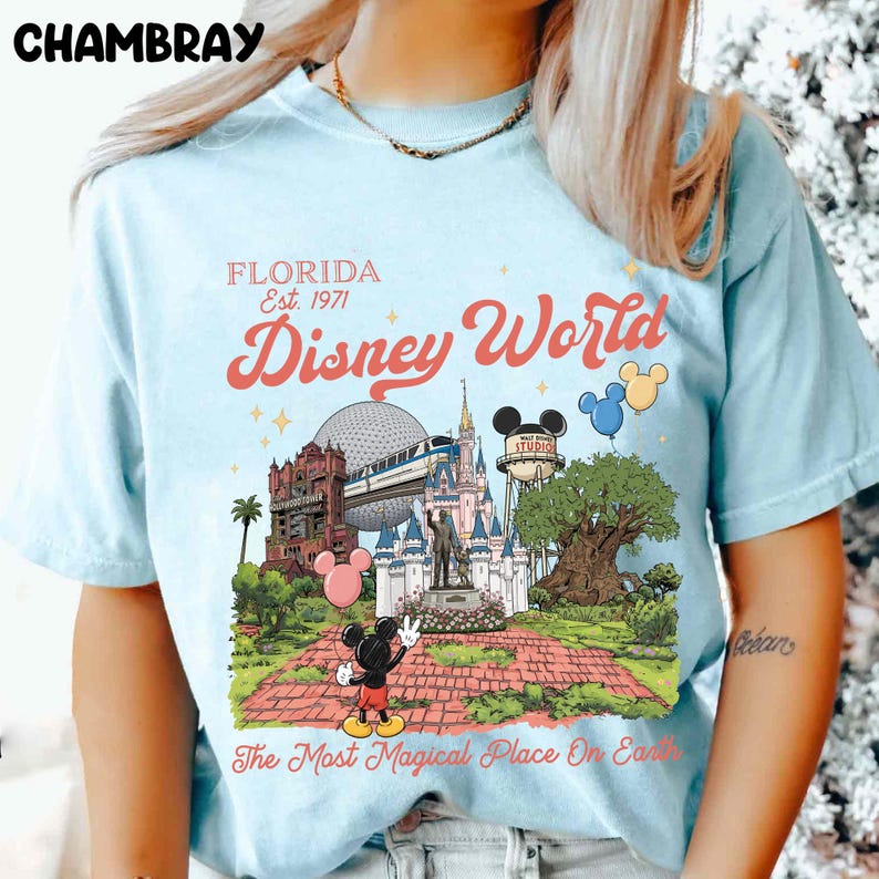 Puede incluir: Camiseta azul claro con un gr&aacute;fico colorido de Disney World. El dise&ntilde;o incluye el texto "FLORIDA Est. 1971" y "Disney World", junto con ilustraciones de caracter&iacute;sticas ic&oacute;nicas del parque y Mickey Mouse. La camiseta tambi&eacute;n tiene el texto "The Most Magical Place On Earth".