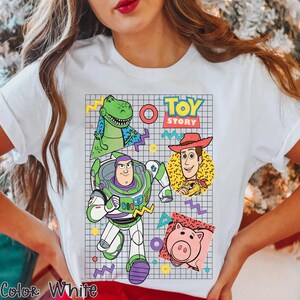 May include: White t-shirt featuring a colorful graphic of characters from the animated film "Toy Story." The design includes Buzz Lightyear, Woody, Rex, and Hamm, set against a grid background with geometric shapes and the words "Toy Story."