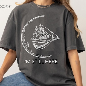 May include: A dark gray t-shirt with a white graphic of a ship sailing on a crescent moon. The text "I'm Still Here" is printed below the graphic.