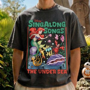 Pu&ograve; includere: T-shirt grigio scuro con una grafica colorata de "La Sirenetta" di Disney. Il design include Ariel, varie creature marine e il testo "Singalong Songs The Under Sea."