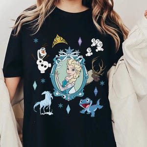 Puede incluir: Camiseta negra con un estampado gr&aacute;fico de Elsa de Frozen. El gr&aacute;fico presenta a Elsa con un vestido azul con una corona y copos de nieve. Otros personajes de Frozen tambi&eacute;n aparecen en el gr&aacute;fico, incluidos Olaf, Sven y los mu&ntilde;ecos de nieve.