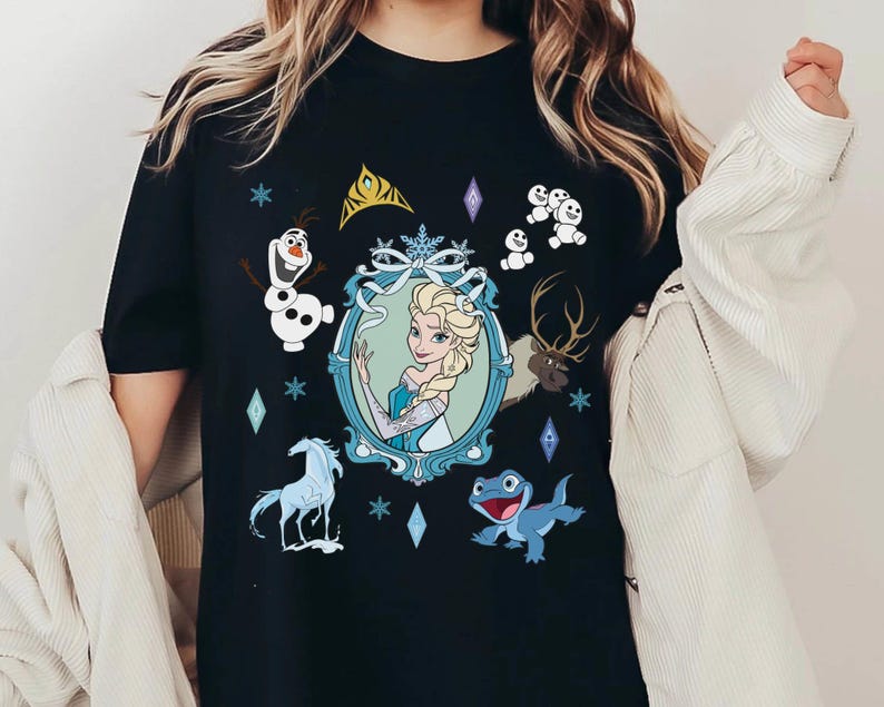 Puede incluir: Camiseta negra con un estampado gr&aacute;fico de Elsa de Frozen. El gr&aacute;fico presenta a Elsa con un vestido azul con una corona y copos de nieve. Otros personajes de Frozen tambi&eacute;n aparecen en el gr&aacute;fico, incluidos Olaf, Sven y los mu&ntilde;ecos de nieve.